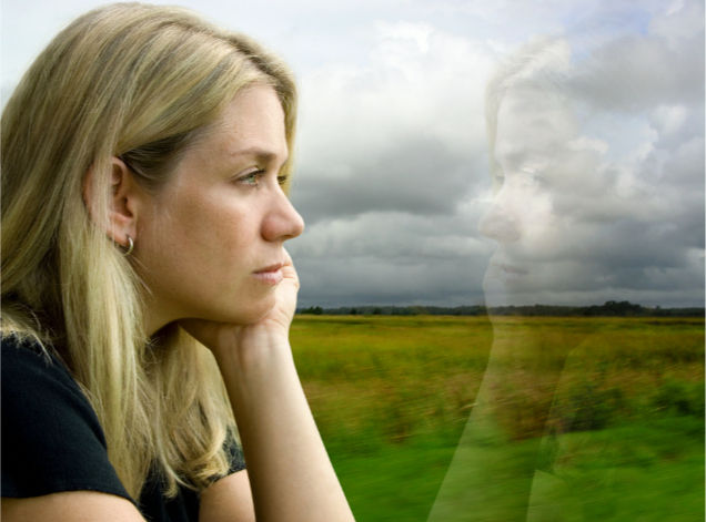 A pensive woman looking out a window, her reflection visible in the glass, a green field with grey clouds stretching beyond her.
