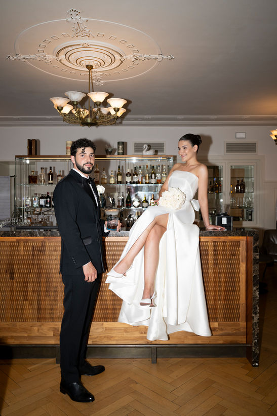 bride is sitting on the bar counter with a drink in her hand, legs crossed. The groom standing next to her.