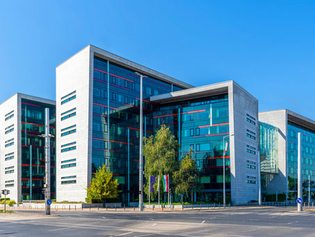 Aréna Corner, modern üveg irodaház, szürke és piros részletekkel, zöld fák előtt. Kék ég és napfény. Úttest az előtérben átkelőhelyekkel.