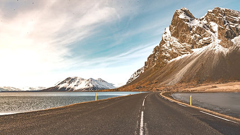 Route goudronnée bordée par l'eau et les montagnes enneigées sous le ciel bleu.