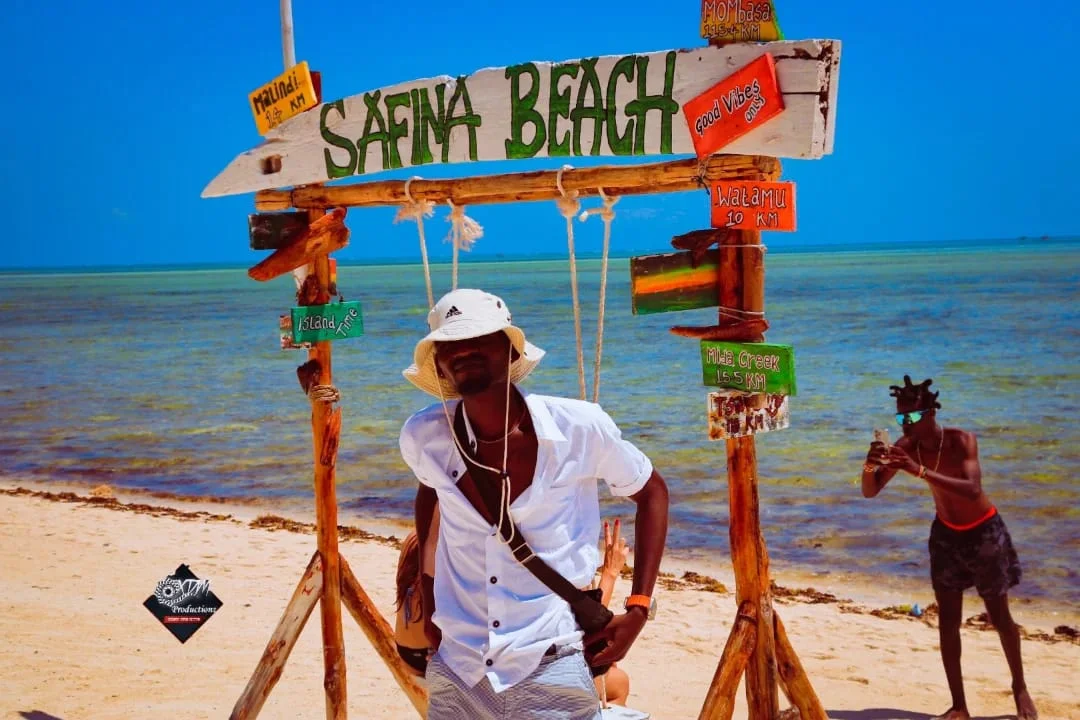 Man in a white bucket hat and shirt poses under a wooden Safina Beach sign with colorful distance markers, on a sunny beach with clear turquoise water