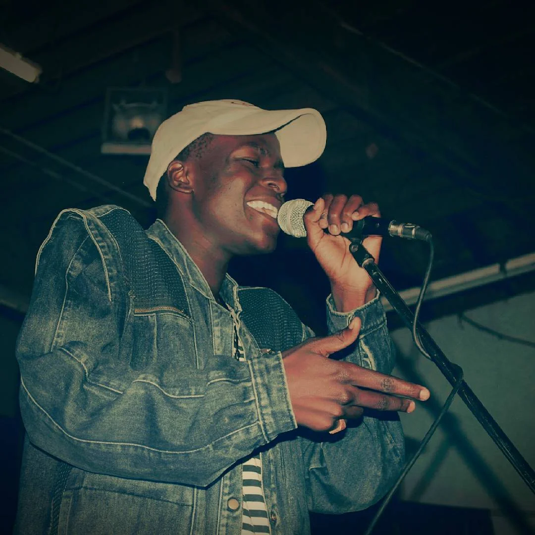 Man in a denim jacket and white cap singing into a microphone on stage, gesturing with his hand.
