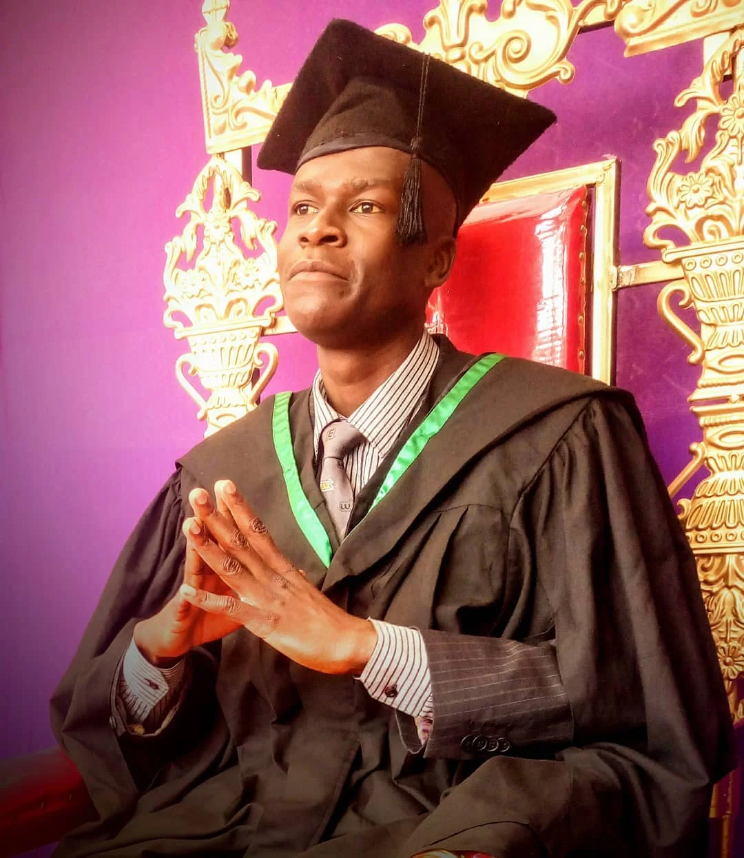 Graduate in black gown and cap with green trim, seated on an ornate gold and red chair against a purple background, hands clasped.