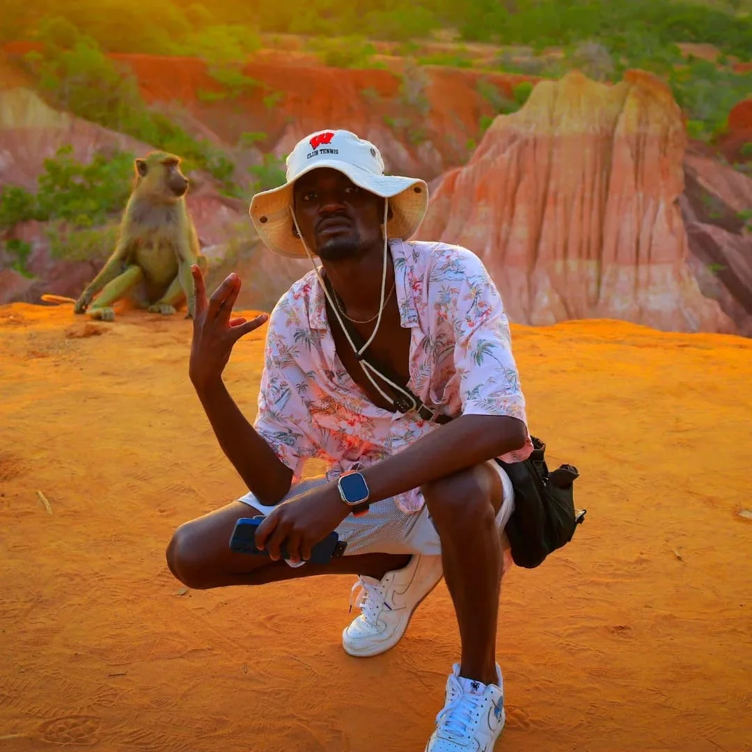 Man in a bucket hat and floral shirt squatting on orange soil with a baboon sitting behind him, against colorful eroded cliffs at golden hour.