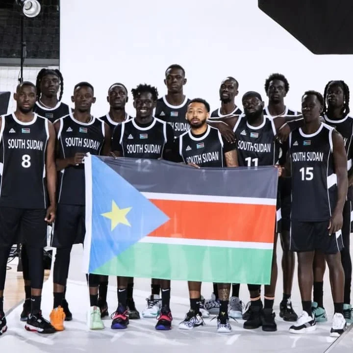 South Sudan basketball team posing together in black jerseys while holding their national flag during a studio photoshoot.
