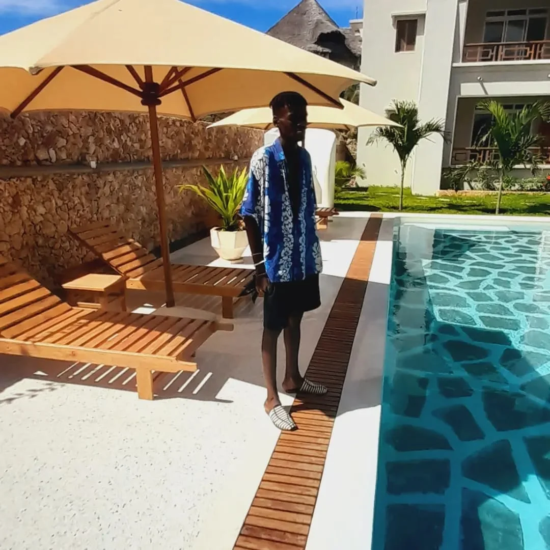 Man in a blue patterned shirt and striped slides stands by a resort pool with wooden loungers and beige umbrellas, next to a stone wall and white building.