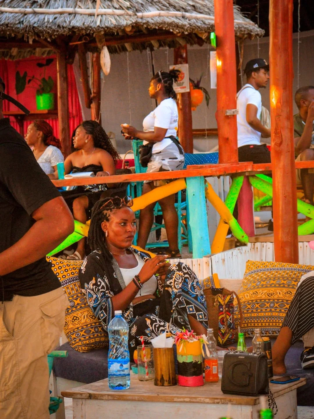Woman with locs in a patterned outfit sits on colorful cushions at a lively outdoor beach bar with thatched roofs, drinks and bottles on the table, other people socializing around her.