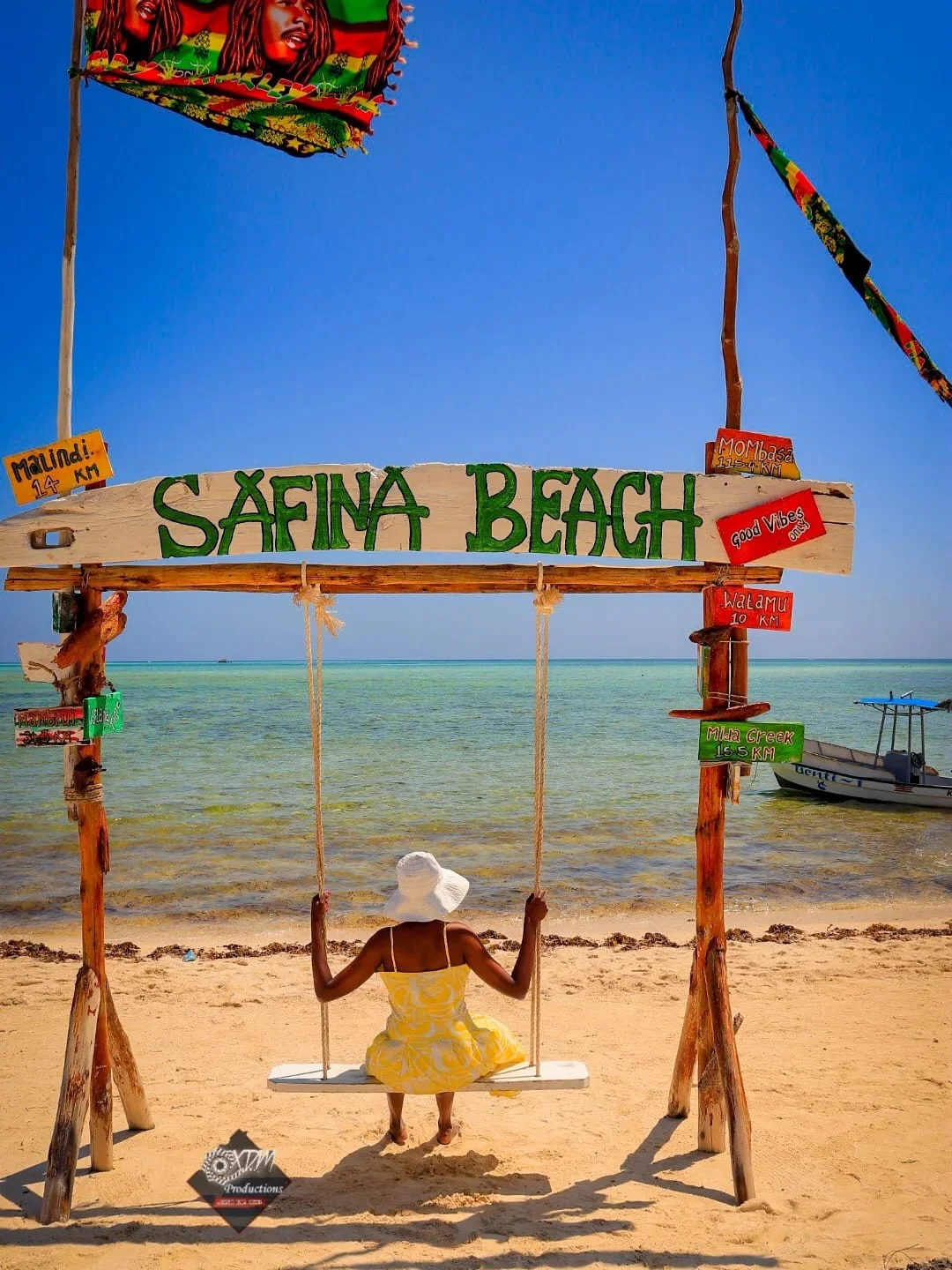 A woman in a yellow dress and white sun hat sits on a beach swing facing the ocean under a wooden sign that reads SAFINA BEACH.