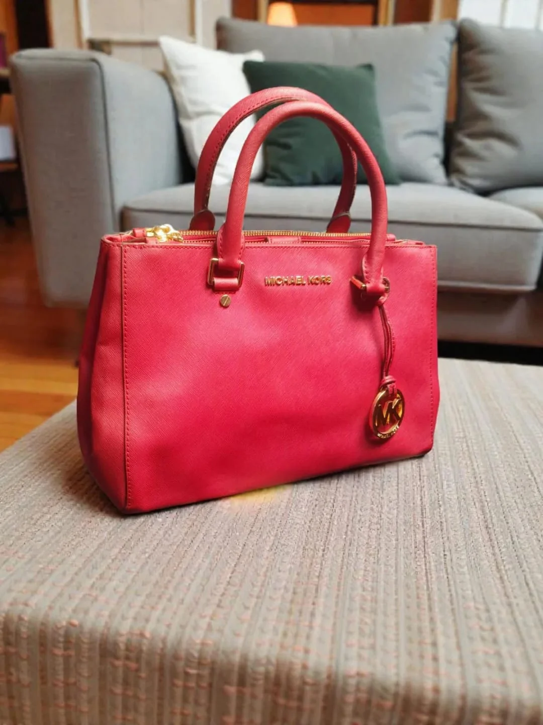 Red Michael Kors leather satchel with gold hardware and MK charm, placed on a textured ottoman in a living room.