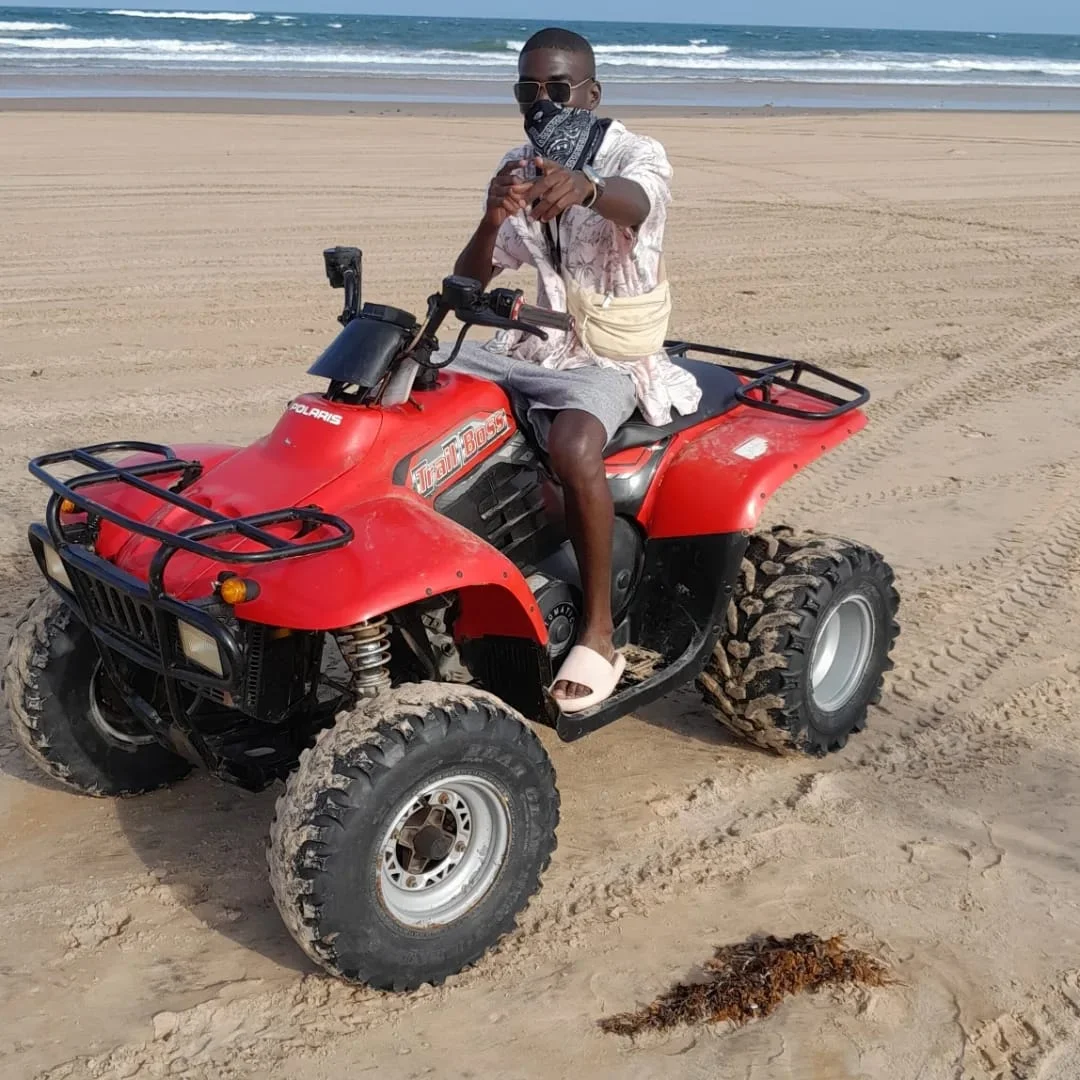 A man wearing sunglasses and a bandana sits on a red Polaris ATV on a sandy beach, pointing toward the camera with the ocean in the background.