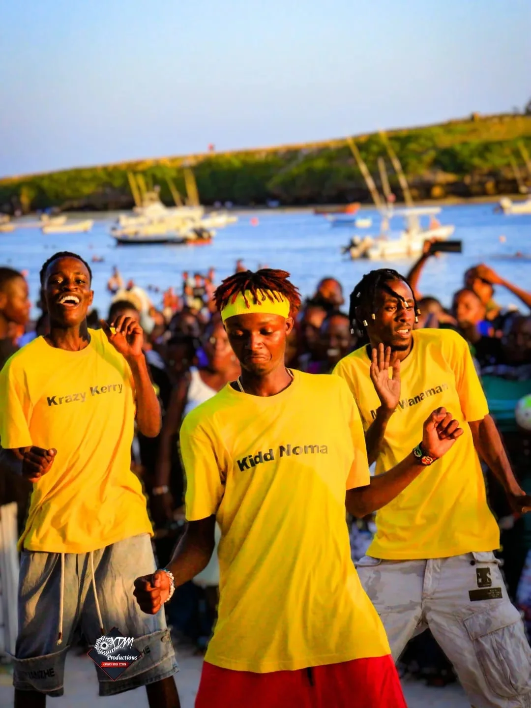 Three young men in matching yellow shirts dance energetically in front of a crowd by the water at sunset, with boats and a shoreline in the background.