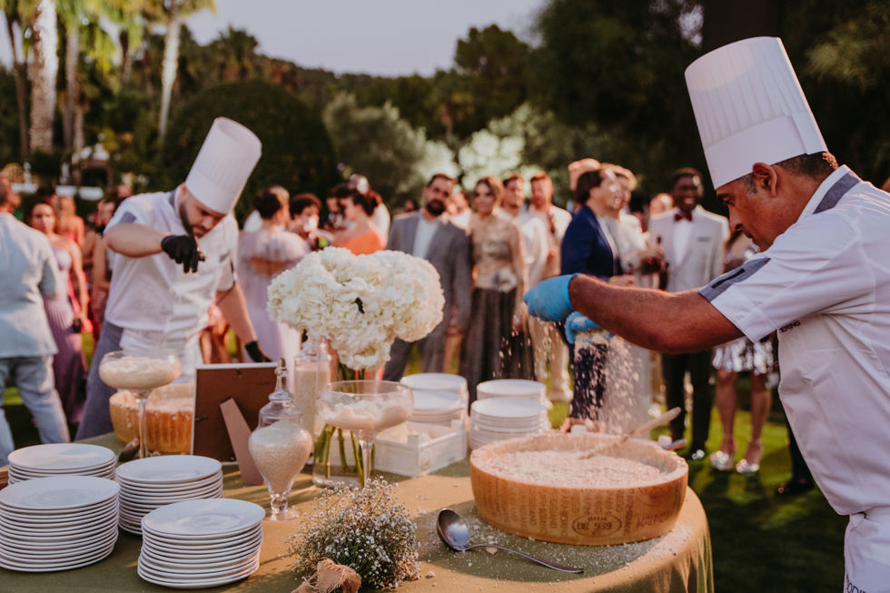 Chefs preparing pasta with a large cheese wheel during a Mediterranean cocktail hour show at an Ibiza wedding, with guests watching in the background. ibiza wedding entertainment. Interactive pasta-making demonstrations, which add a sense of entertainment, allowing guests to witness the artistry behind creating homemade pasta.