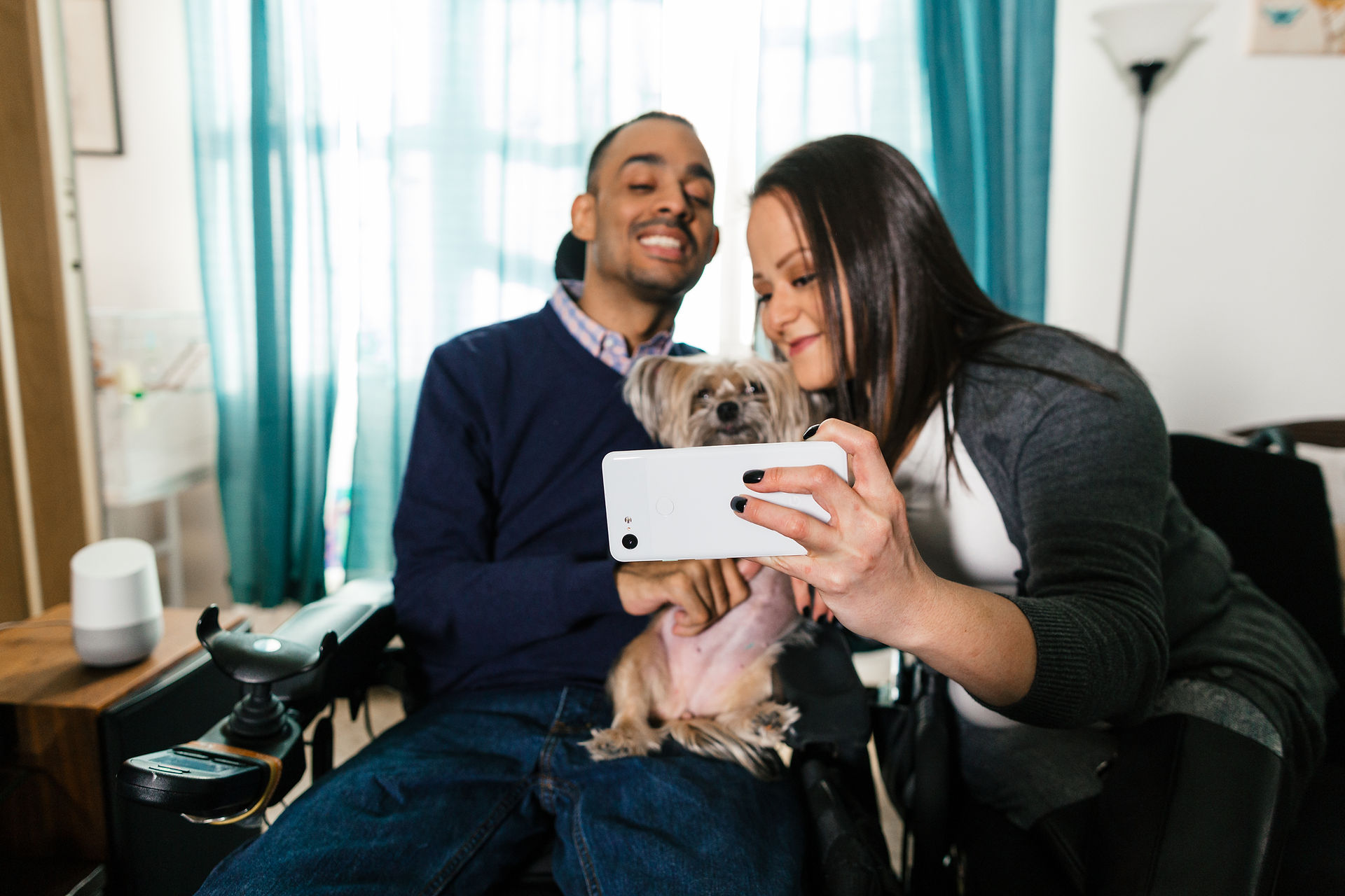 A man sits in a wheelchair and a woman sits next to him while holding a yorkie terrier. Both pose for a selfie with a Google Pixel 6. Image courtesy Google.