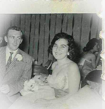Photo of my Aunt at her prom. She is a young, white woman with a round face, and dark curly hair to her chin. She is wearing a white strapless gown and a large, sparkling earring peeking out from behind her hair on one side. She is seated in a chair and looking straight at the camera and smiling. A table is to the left of her and a young, white man with slicked back hair, seated on the other side, is wearing vertical striped suit with a tie and corsage. He is staring at my Aunt and smiling softly. In the background are metal folding chairs, people talking, and streamers on the walls. The photo is black and white.