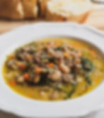 A wide, white rimmed bowl is filled with a creamy looking soup. The ingredients are heaped in the middle of the bowl and surrounded by an opaque, golden broth. You can see white beans, carrots, onions, greens, sausage, and some small pieces of herbs. In the background, there is an out of focus wooden cutting board with a loaf of sliced bread on it .
