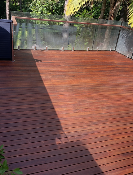 Freshly restored timber deck with clean, even stain finish, cleared surface and trimmed surrounding greenery, creating a neat and well-presented outdoor area with glass balustrade.