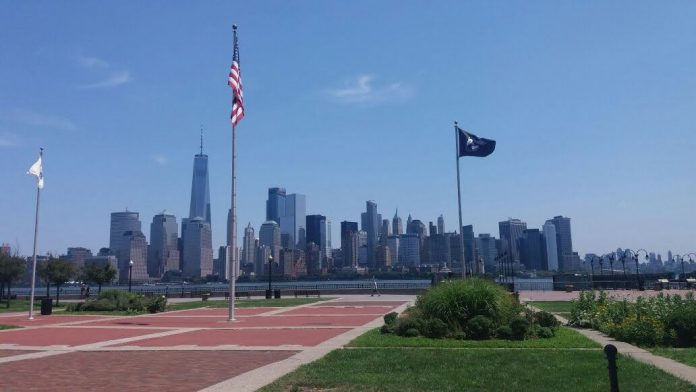 Skyline of Manhattan with flags waving in a park. Clear blue sky, red brick paths, and lush green grass set a peaceful mood.