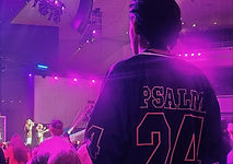 a young man stands in a church pew wearing a psalms 24 jersey style shirt with vibrant florescent lights in the background