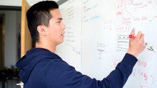 A man in a hoodie writing on a whiteboard with a red whiteboard marker.