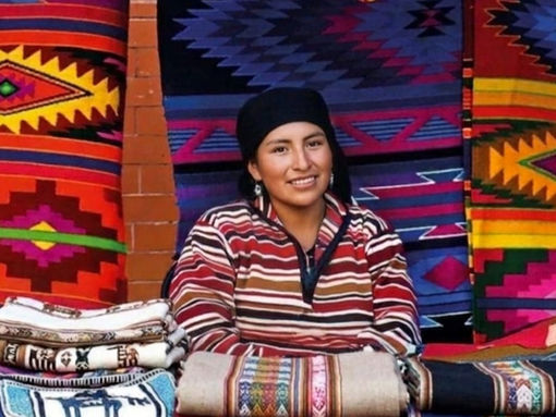 Woman wearing a striped Andean textile in front of colorful handwoven blankets and rugs