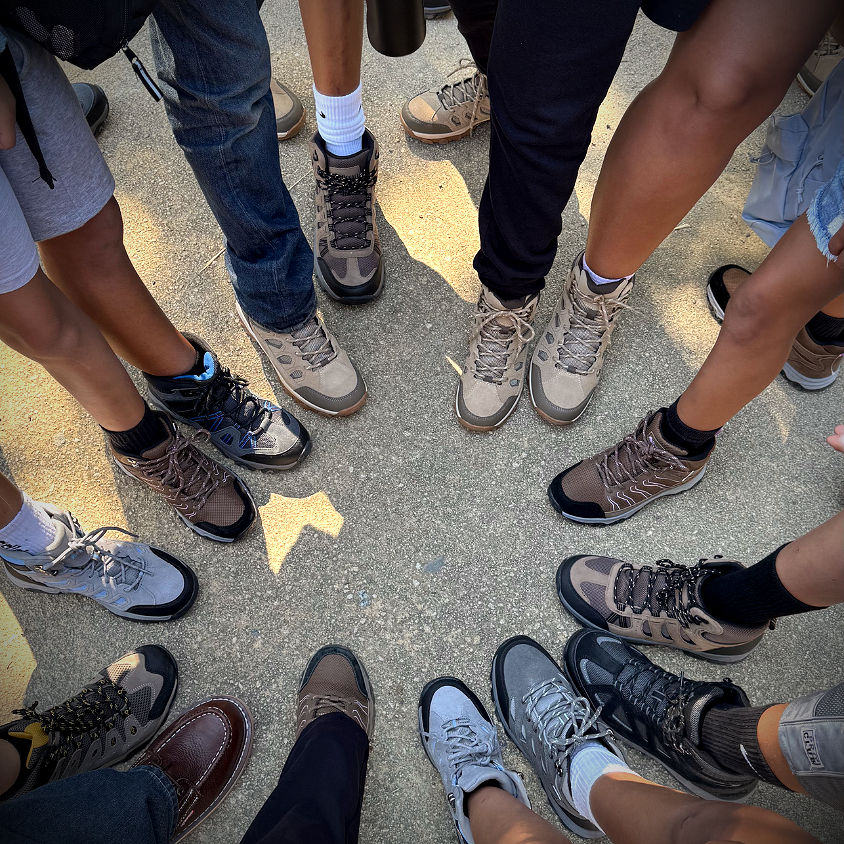 Circle of diverse youth proudly showing off new hiking boots during field trip