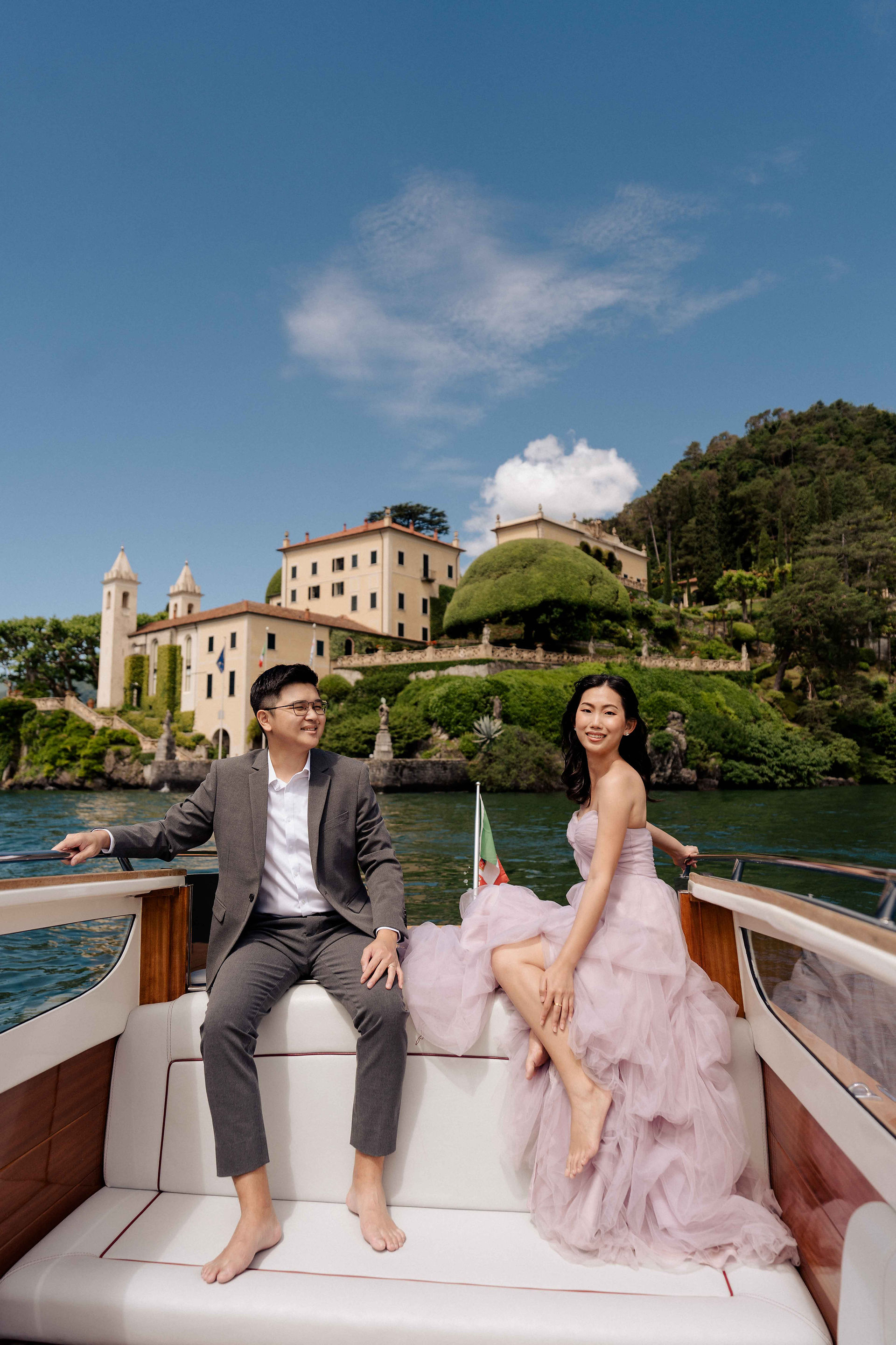 Couple in wedding attire posing on their private boat with stunning view of Villa Balbianello behind
