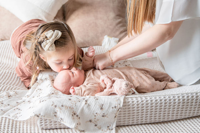 Een moeder kleedt haar baby aan terwijl haar zus haar een kusje op haar wang geeft. 