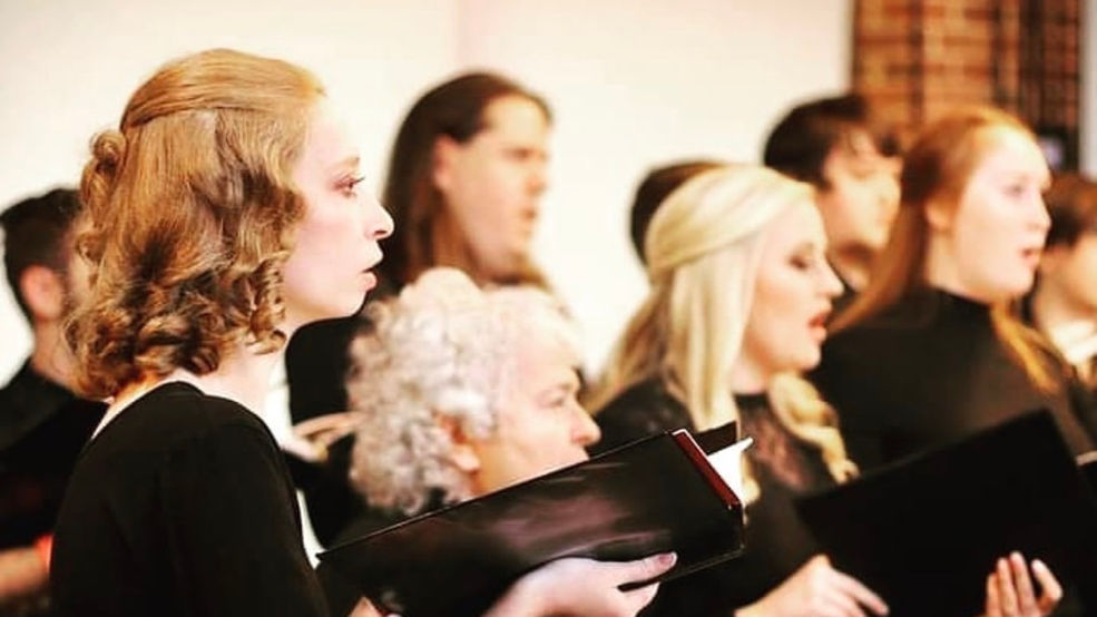 A choir pictured from the side sings while dressed in all black and holding books of music.