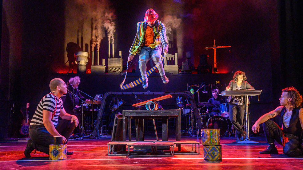 Performers and musicians on a red lit stage watch a man in construction gear jumprope on a small teeter totter.