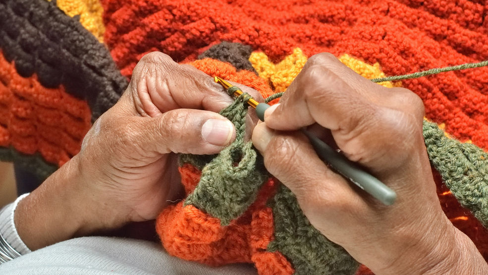 Photo of an older woman's hands from above as she knits a blanket using green, orange, brown, and yellow yarn.