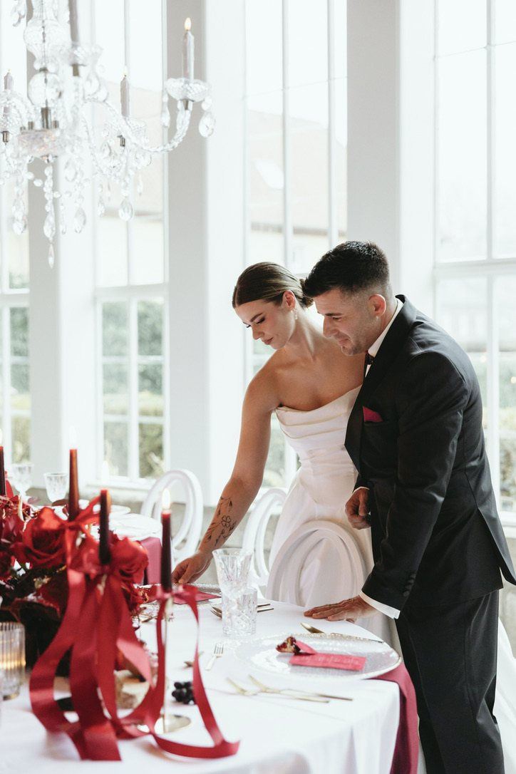 Brautpaar an der Hochzeitstafel mit roten Rosen, Anthurien und Satinbändern in der Orangerie Gut Ostenwalde, Hochzeitsfloristik von Ivy & Rose