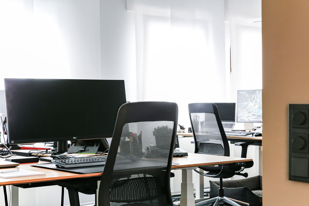 Schreibtische in den Büroräumen der Bodensteiner Gruppe. Je zwei Schreibtische sind gegenübergestellt, ein weiterer steht am Kopf der beiden. Jeder Arbeitsplatz ist mit einem schwarzen Bürostuhl und einem großen schwarzen Monitor ausgestattet.
