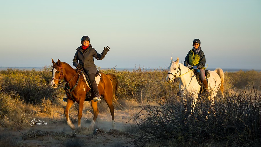 Dr Z waves to the camera as she and a friend ride horses on a sandy beach with the ocean in the background.