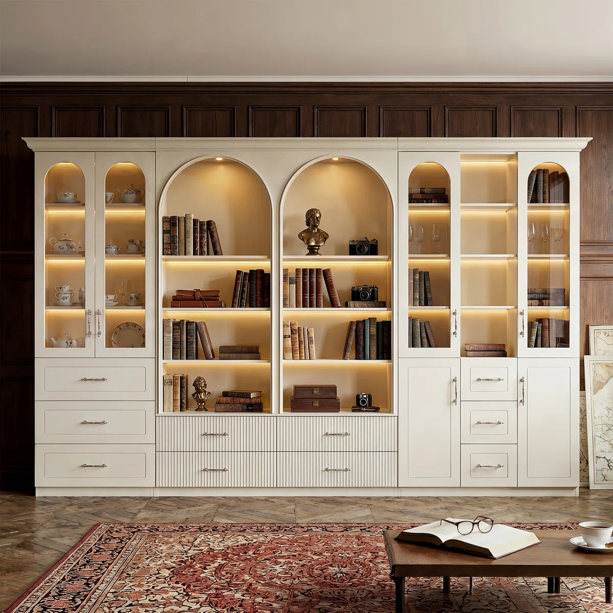 Custom white built-in bookcase wall with arched shelves, warm LED lighting, glass-front cabinets, and curated book styling. A Nancy Meyers-inspired home library showcasing quiet luxury interiors, classic millwork, timeless design, and elegant traditional decor.