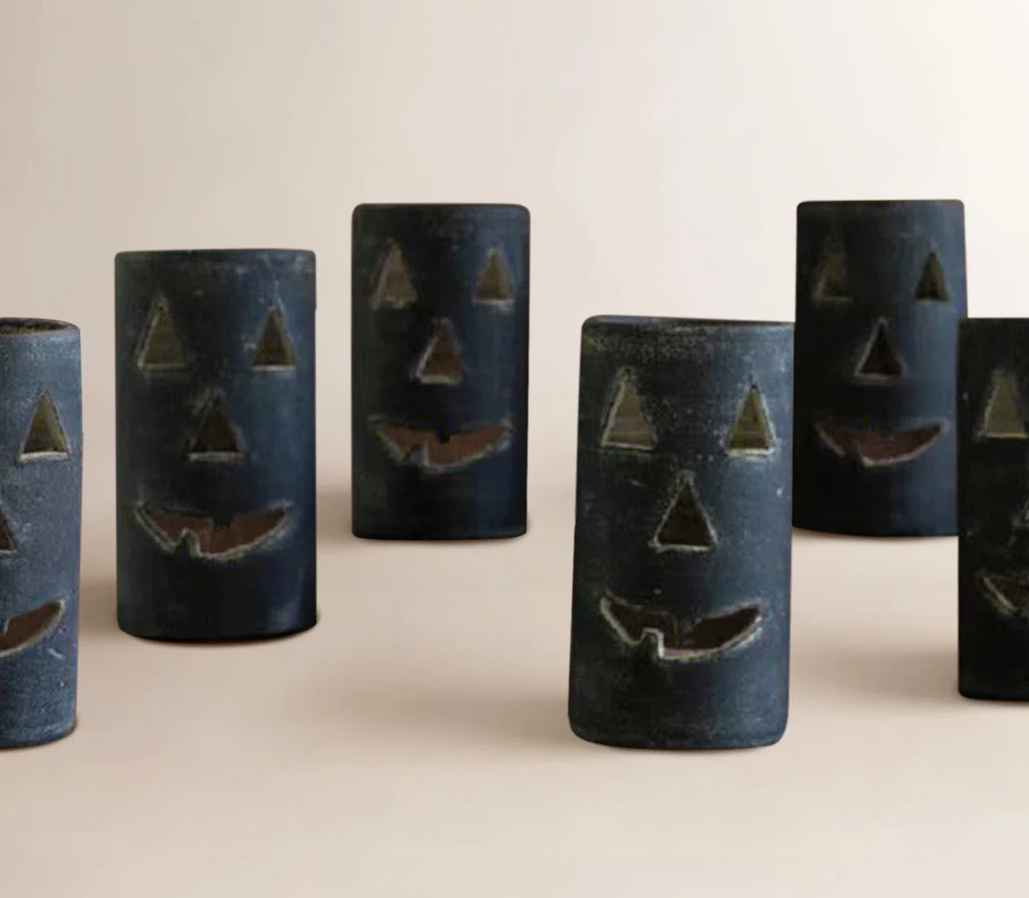 Five black cylindrical jack-o'-lantern candles with carved faces on a plain beige background. Esmeray Clay Tealight Candle Holder