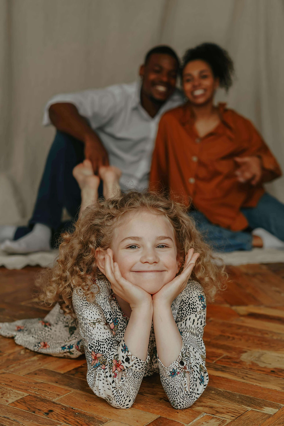 A girl with curly hair smiles, resting chin on her hands, lying on wooden floor with two smiling adults sit blurred in the background | parent coach in Boulder County, CO | parenting coach online | parent coaching services | Arapahoe | Foothills | Boulder