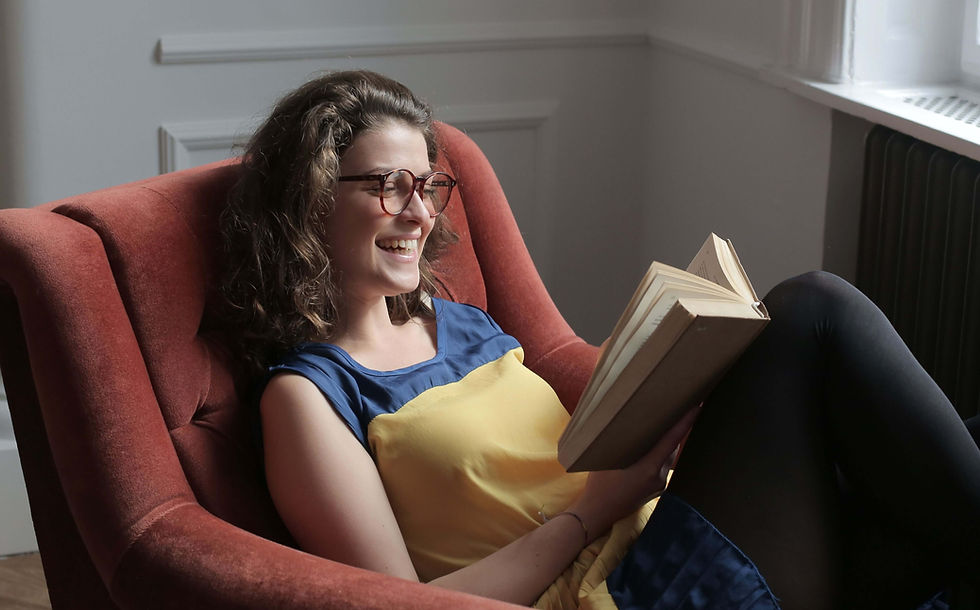 Smiling woman in glasses reads a book in a rust-red armchair by a window in a bright room | parent coaching therapy in Boulder County, CO | parent coaching services | feeling overwhelmed as a parent | Superior | Lafayette | North Boulder