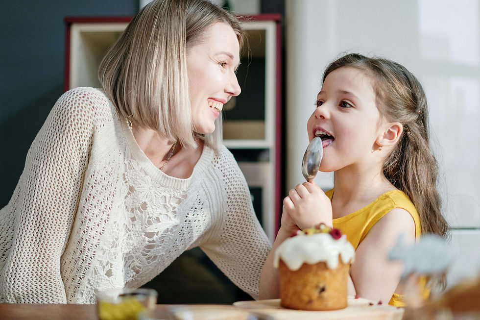 A woman smiles at a girl licking a spoon, with a decorated cake nearby on a sunny kitchen table | parent coach in Boulder County, CO | parenting coach online | parent coach | Arapahoe | Foothills | Boulder