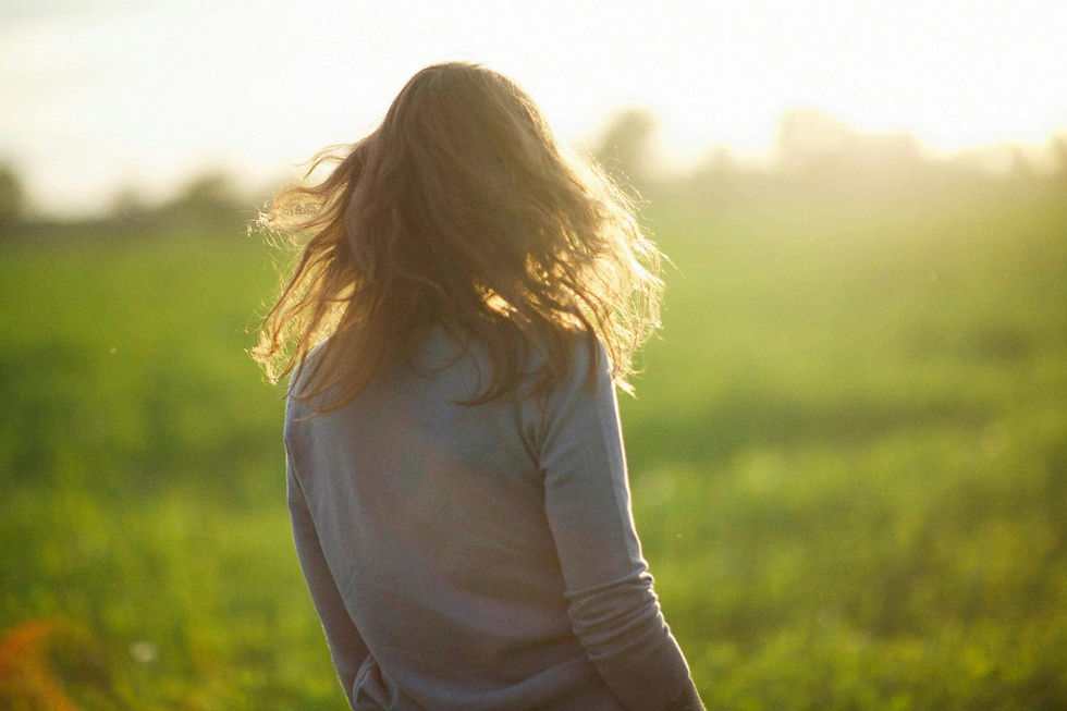 A woman stands in a sunlit green field with her back to the camera, her hair blowing in the wind, representing reflection and healing as explored by a South Asian therapist in NYC.
