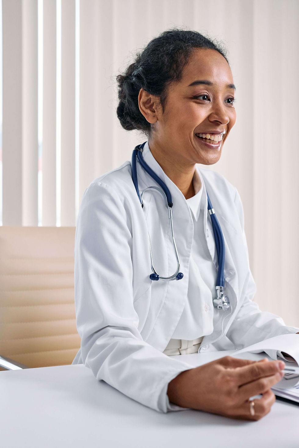 Smiling doctor in a white coat with a stethoscope sits at a desk in a bright office with vertical blinds | anxiety therapy for professionals in Manhattan, NY | therapist for anxiety | high functioning anxiety | Upper West Side | Upper East Side | Financial District