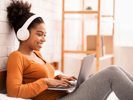 A young woman smiles while using a laptop with headphones on, comfortably settled at home — reflecting the accessible, welcoming nature of online ketamine therapy in Colorado. For those exploring ketamine-assisted therapy in Colorado, the process often begins with exactly this kind of relaxed, at-home telehealth experience. Compass Healing Project makes it easy to take that first step toward healing from wherever you are in the state.
