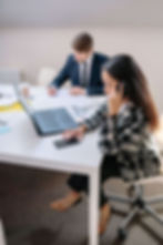 Woman on phone and laptop at white office desk while man in suit writes in background, focused work scene | anxiety therapy in Manhattan, NY | high functioning anxiety | therapist for anxiety | Midtown | Tribeca | Gramercy Park