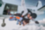 A lady in blue jumpsuit attached to man in white jumpsuit jumping out of a plane