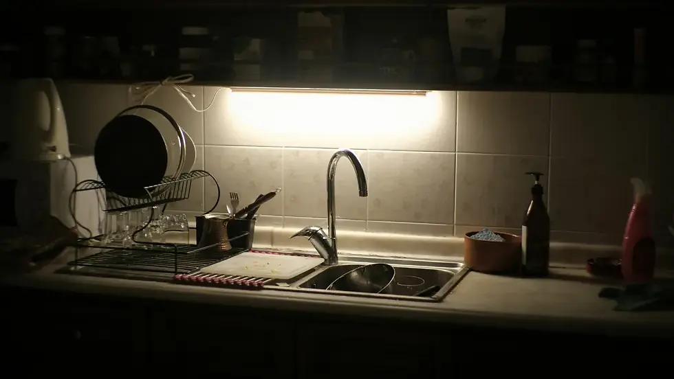 Dim kitchen sink with dishes drying on a rack under a bright cabinet light, bottles on the counter, quiet and messy.