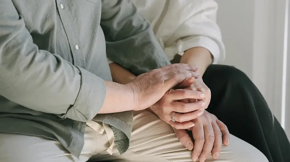 Two people sit closely, hands gently clasped in a comforting gesture. They wear neutral-colored clothing, conveying a serene and supportive mood.