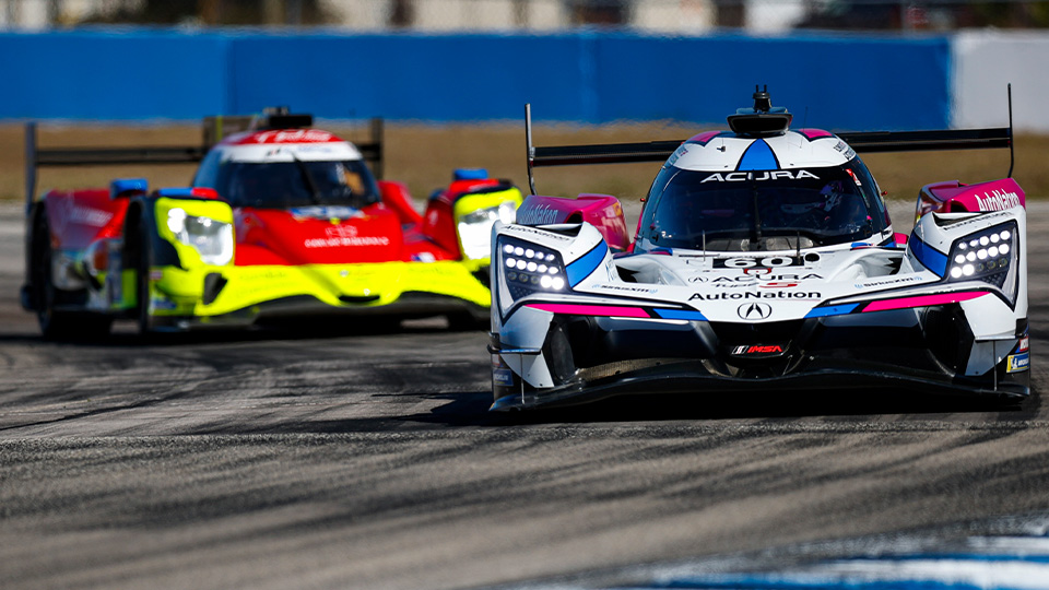 Acura GTP race car leads a neon yellow-red prototype on track, headlights on, with Acura and AutoNation text against a blue wall for IMSA Weathertech Championship at Indianapolis Motor Speedway.
