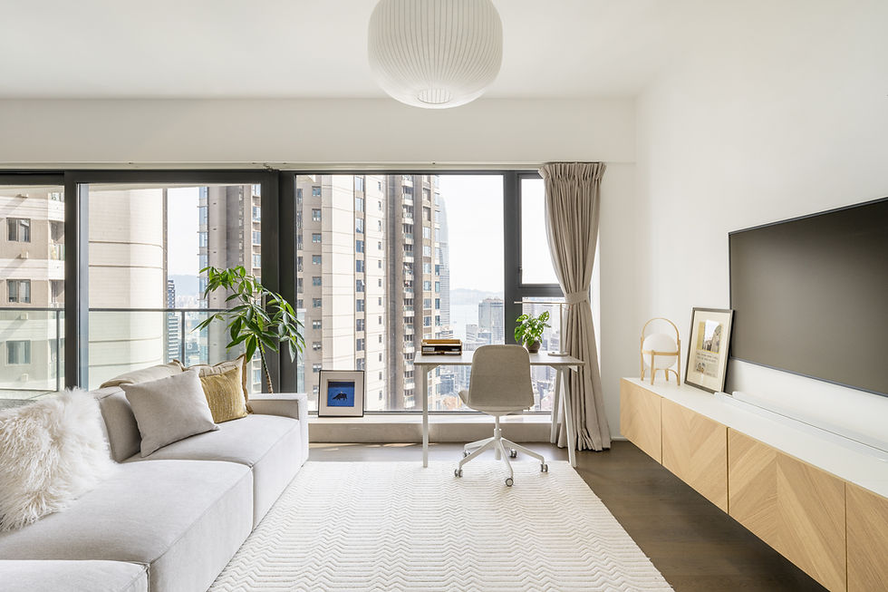 Modern living room with gray sofa, desk, chair, and TV. Bright windows show cityscape. Neutral tones, plant on desk, cozy atmosphere.