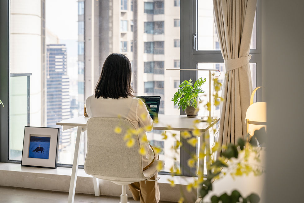 A woman sits at a desk with a laptop, in a bright room with large windows showing city buildings. A plant and framed art are nearby.
