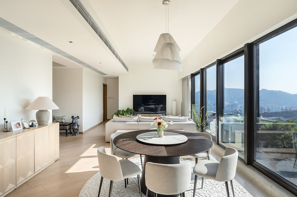 Modern living room with round dining table, beige chairs, and a large TV. Floor-to-ceiling windows offer city views. Bright and airy.
