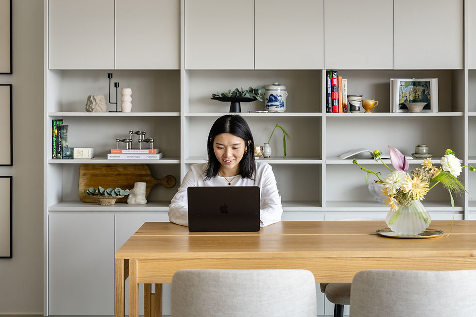Woman smiling at a laptop at a wooden table. Behind her, a bookshelf with books, decor, and flowers. Bright and tidy room.